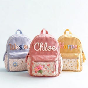 May include: Three corduroy backpacks in light blue, pink, and yellow. Each bag features a front zippered pocket with a floral quilted pattern and a personalized name embroidered in large, raised script letters. A small crocheted flower is attached.