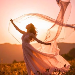 Op de afbeelding: Een vrouw in een vloeiende, lichtroze jurk staat in een veld met hoog gras, met uitgestrekte armen, met een doorschijnende stof die in de wind wappert. De ondergaande zon werpt een warme, gouden gloed over de sc&egrave;ne, waardoor een romantische en etherische sfeer ontstaat.
