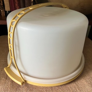 May include: A white plastic cake carrier with a yellow handle and base. The carrier has a rounded shape and a lid that fits snugly on the base. The handle is arched and has circular cutouts. The carrier is designed to transport and store cakes.