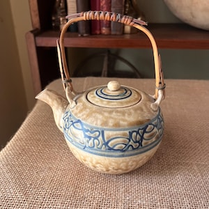 May include: A round ceramic teapot with a tan, mottled glaze and a blue hand-painted swirling pattern around the center. It features a matching lid with a small knob and a curved spout. A flexible, arched handle made of wrapped bamboo is attached.