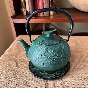 May include: A dark green cast iron teapot featuring a textured surface with embossed monkey figures and foliage. The teapot has a black arched handle, a matching lid with a leaf-shaped knob, and sits on a circular green coaster with a wave pattern.