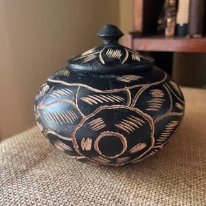 May include: A round black lidded container featuring carved floral patterns with light-colored wood grain exposed within the etched lines. The lid has a small pointed knob at the top, and the body displays a repeating circular motif.