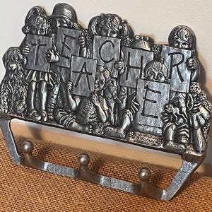 Può includere: Un portachiavi in metallo color argento con bambini che tengono lettere che compongono la parola "TEACHER". Il portachiavi ha tre ganci ed è decorato con figure dettagliate di bambini. Un regalo premuroso per gli educatori.