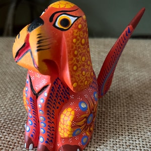 May include: A vibrant, hand-painted wooden dog figurine. The folk art sculpture features a bright orange base with yellow and blue floral and dot patterns. The dog has a long tail and is posed in a sitting position.