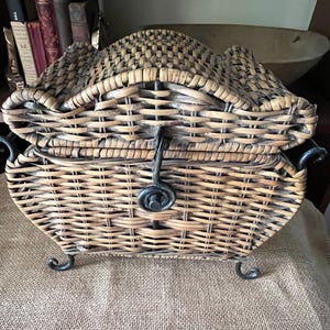 May include: Woven wicker basket with a curved lid and metal accents. The basket has a rectangular shape with a decorative metal clasp and feet. The basket is made of light brown wicker and is likely used for storage or decoration.