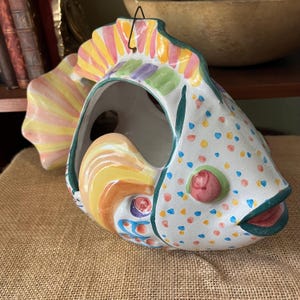 May include: A ceramic fish-shaped wall pocket or planter featuring a white base with multicolored polka dots, a yellow and pink striped tail, and a large open center. The fish has a raised pink eye, a dark green outline, and a metal hanging wire.