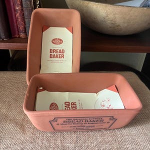 May include: Two rectangular terracotta bread baker dishes by Henry Watson's Pottery. Each dish features a light brown clay finish and a paper insert with the text "Henry Watson's Pottery Limited" and "Bread Baker" printed in red and black ink.