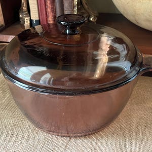 Puede incluir: Una fuente para horno redonda de vidrio marrón vintage con tapa a juego. El plato tiene dos asas laterales y un pomo en la tapa. El vidrio tiene un ligero tinte y es reflectante. La fuente para horno está sobre una superficie beige.