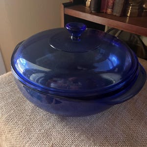 May include: A deep cobalt blue glass casserole dish with a matching lid. The lid features a central knob handle surrounded by a circular, radiating ribbed pattern. The bowl has integrated side handles and a smooth, translucent finish.