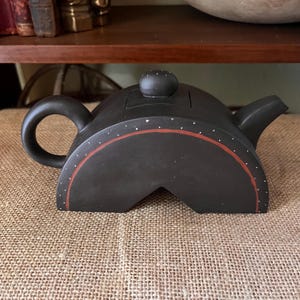 May include: A matte black ceramic teapot with a semi-circular, arch-shaped body. It features a curved handle, a short spout, and a small lid with a knob. The design includes a thin, hand-painted reddish-brown arc and small white dots.
