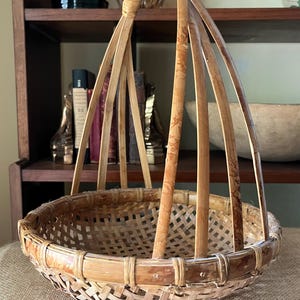 May include: A handcrafted, round basket with a woven base and a tall, arched handle. The basket is made of light brown bamboo and features a lattice weave design. The handle is supported by vertical bamboo rods. The basket is sitting on a burlap surface.