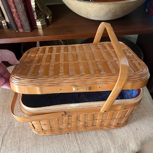 May include: A rectangular woven light brown wooden basket with a hinged lid and a single arched handle. The interior is lined with a dark blue fabric featuring a subtle repeating paisley pattern. The basket has a flat base and rounded corners.