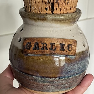 May include: A ceramic garlic keeper with a cork stopper. The jar is a mix of colors, including white, brown, and blue. The word "GARLIC" is printed on a wooden plaque attached to the jar. The jar has several small holes.
