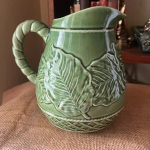 May include: A sage green ceramic pitcher featuring a raised relief pattern of large leaves on the body and a rope-style handle. The base of the pitcher is decorated with a textured lattice design, and a rope-like border encircles the top rim.