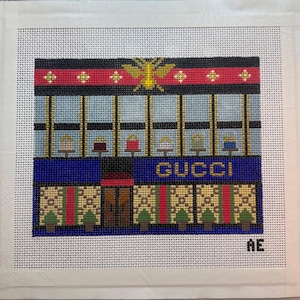 May include: Needlepoint canvas featuring a stylized depiction of a Gucci store. The design includes the brand's name in gold lettering, a blue facade, and a red and gold striped awning. The background is a light blue color.