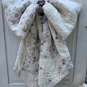 May include: A large decorative bow crafted from quilted fabric featuring a floral pattern with yellow and purple roses on an off-white background. The center of the bow is secured with a small, dark, flower-shaped embellishment and a ring.