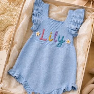 May include: A light blue knit romper featuring ruffled shoulder straps and a ruffled hem. The chest displays the name Lily embroidered in colorful script, flanked by two small white flower appliques. Three buttons are visible at the bottom.