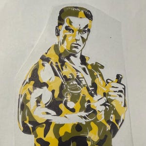 May include: A decal featuring a stylized graphic of a soldier in a yellow, black, and white camouflage pattern. The figure has a stern expression, short hair, and is wearing a tactical jacket with a canteen visible at the chest area.