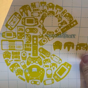 Pode incluir: Folha de adesivos de vinil amarelo com vários designs de controladores de videojogos, dispostos em um padrão circular. Inclui controladores de diferentes consolas de jogos, como Nintendo e Playstation. O fundo é uma grelha.