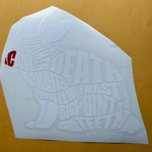 Puede incluir: Calcomanía blanca con forma de conejo con el texto "DEATH WANTS YOU ALL WITH NASTY BIG POINTY TEETH". Una "C" roja está en la esquina superior izquierda. La calcomanía está sobre un fondo amarillo.