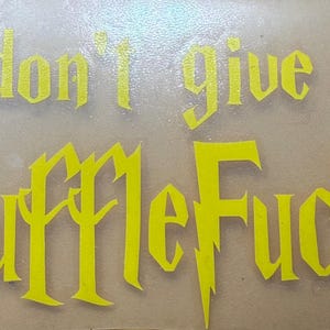 Può includere: Testo giallo su sfondo chiaro con la scritta "I don't give a HuffleFuck" in un carattere gotico. Il testo è probabilmente per un fan di Harry Potter.