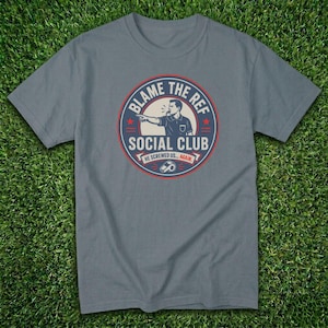 May include: A gray t-shirt with a circular graphic that reads "BLAME THE REF SOCIAL CLUB." The graphic features a referee pointing and the text "HE SCREWED US... AGAIN." The shirt is displayed on a green grassy surface.