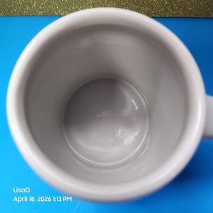 May include: A white ceramic mug with a handle, viewed from above. The interior is a light gray color. The mug is set against a blue background with a gold glittery surface in the background. The text "LisaG April 18, 2026 1:13 PM" is visible.