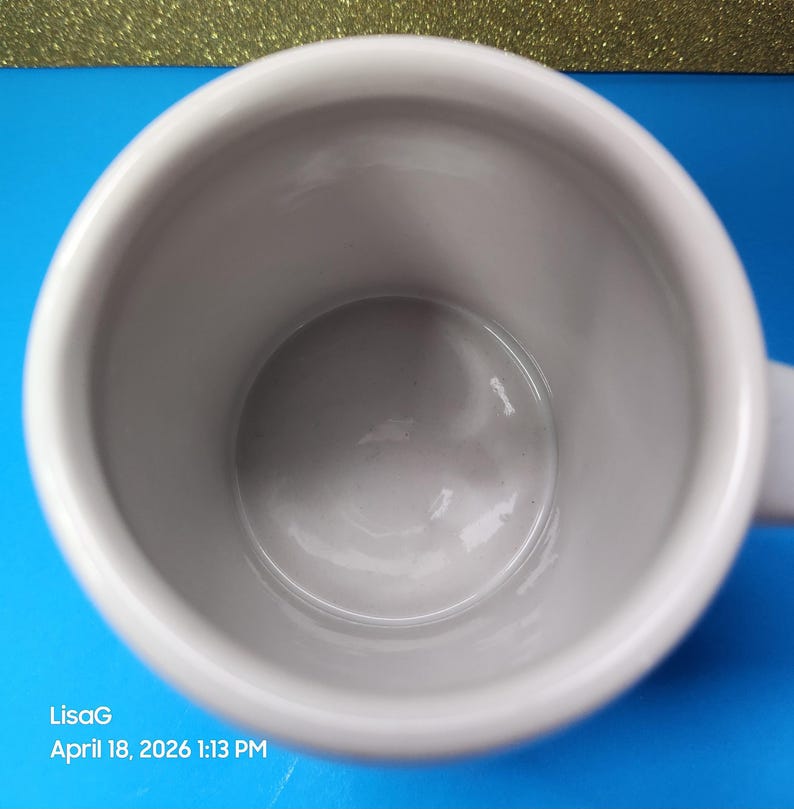 May include: A white ceramic mug with a handle, viewed from above. The interior is a light gray color. The mug is set against a blue background with a gold glittery surface in the background. The text "LisaG April 18, 2026 1:13 PM" is visible.