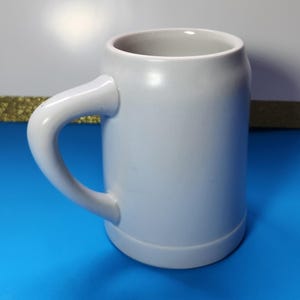 May include: A light gray ceramic beer stein with a curved handle. The mug has a cylindrical body and a slightly flared base. The handle is thick and rounded, and the interior of the mug is a lighter shade of gray. The stein is designed for holding beverages.
