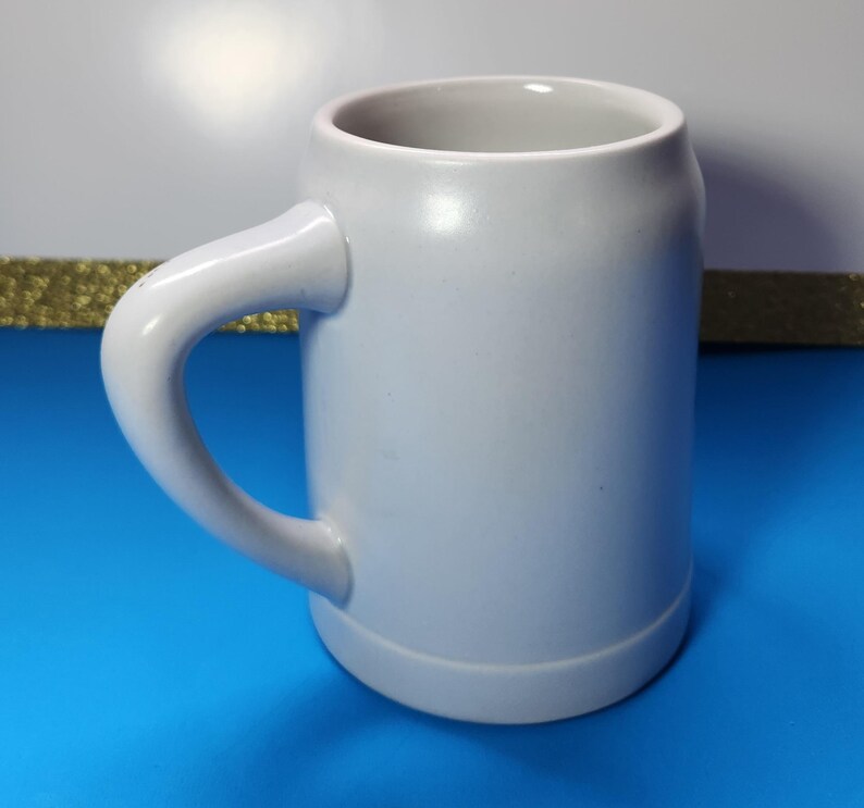 May include: A light gray ceramic beer stein with a curved handle. The mug has a cylindrical body and a slightly flared base. The handle is thick and rounded, and the interior of the mug is a lighter shade of gray. The stein is designed for holding beverages.