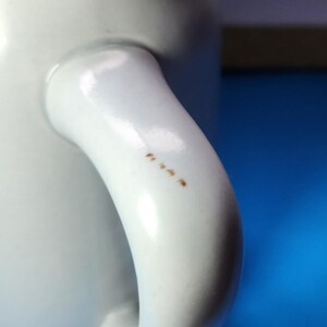 May include: Close-up of a white ceramic mug with a curved handle. The handle has several small brown spots. The mug is set against a blue background.