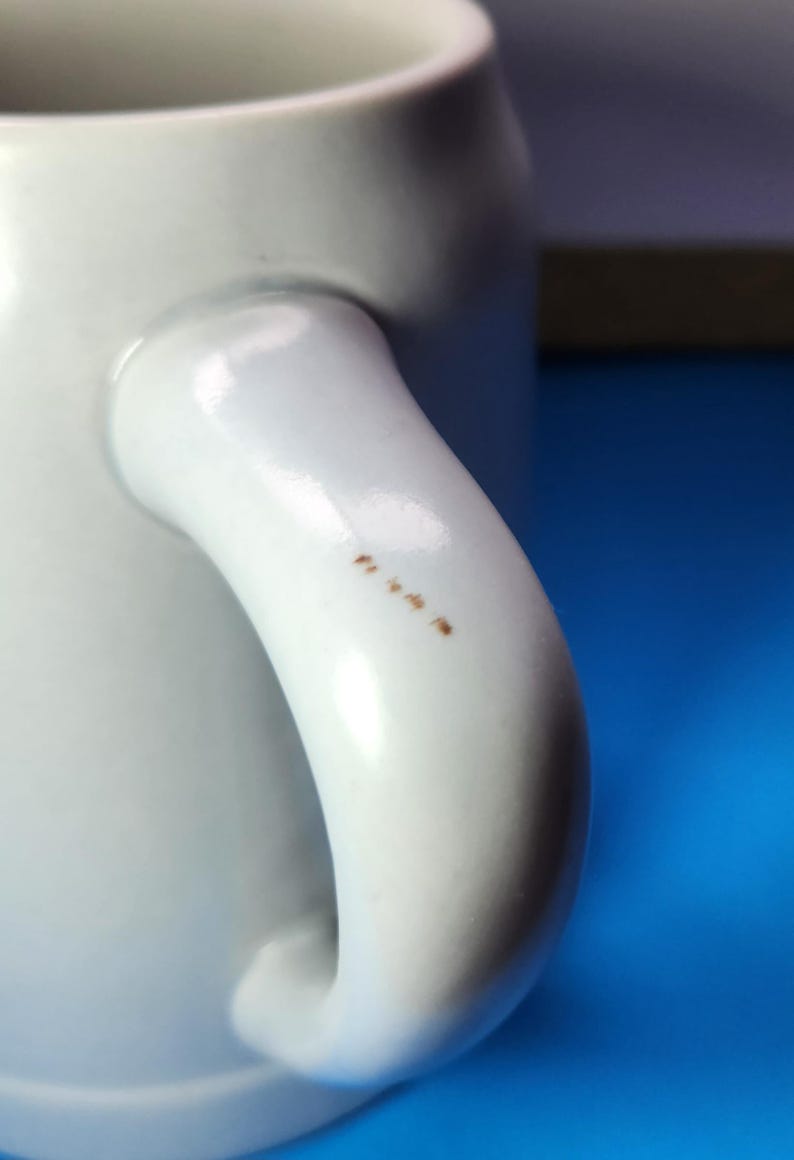 May include: Close-up of a white ceramic mug with a curved handle. The handle has several small brown spots. The mug is set against a blue background.