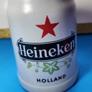 May include: White ceramic beer stein with the Heineken logo, a red star, and green leaf accents. The stein has a cylindrical shape and the word "HOLLAND" is printed at the bottom. A measuring tape is in the foreground.