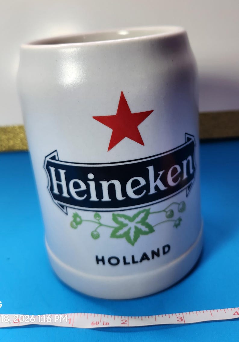 May include: White ceramic beer stein with the Heineken logo, a red star, and green leaf accents. The stein has a cylindrical shape and the word "HOLLAND" is printed at the bottom. A measuring tape is in the foreground.
