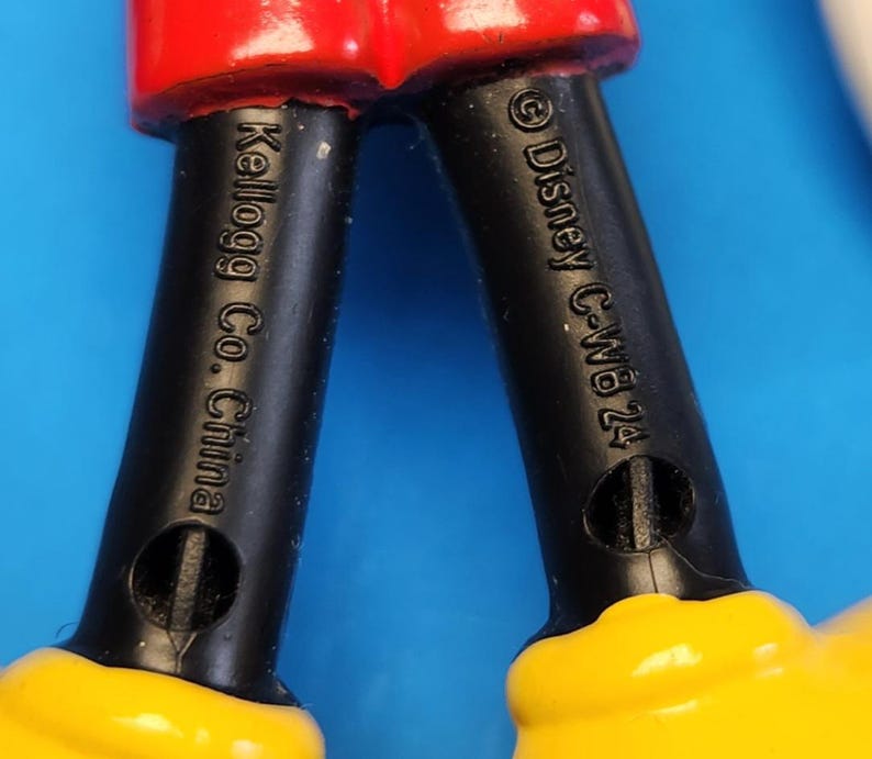May include: Close-up of a toy figure's legs. The legs are black with yellow shoes and red shorts. The text "Kellogg Co. China" and "&copy; Disney C-WB 24" are visible on the legs.