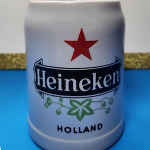 May include: A white ceramic beer stein with the Heineken logo, featuring a red star, a black banner with the brand name, and green leaf accents. The word "HOLLAND" is printed at the bottom.