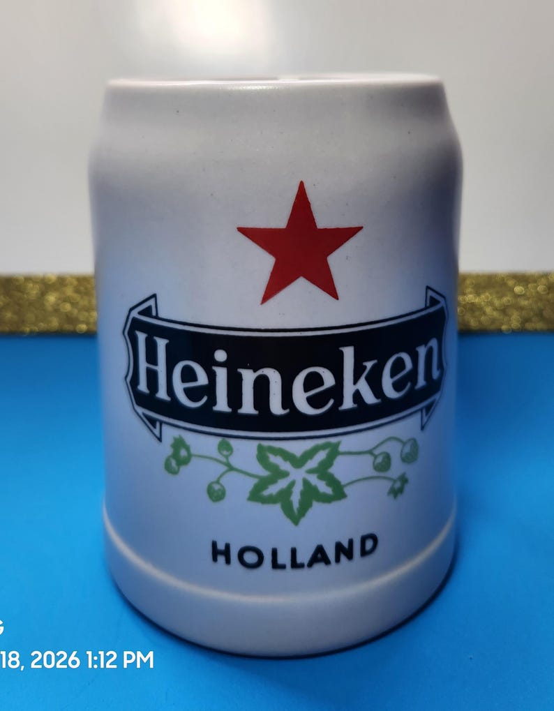 May include: A white ceramic beer stein with the Heineken logo, featuring a red star, a black banner with the brand name, and green leaf accents. The word "HOLLAND" is printed at the bottom.