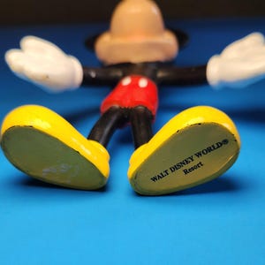 May include: A Mickey Mouse figurine with yellow shoes, red shorts, black arms and legs, white gloves, and a tan head. The words "WALT DISNEY WORLD Resort" are printed on the bottom of the shoes. The figurine is on a blue surface.