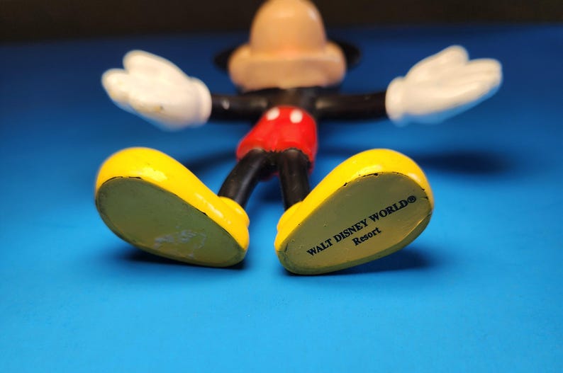 May include: A Mickey Mouse figurine with yellow shoes, red shorts, black arms and legs, white gloves, and a tan head. The words "WALT DISNEY WORLD Resort" are printed on the bottom of the shoes. The figurine is on a blue surface.