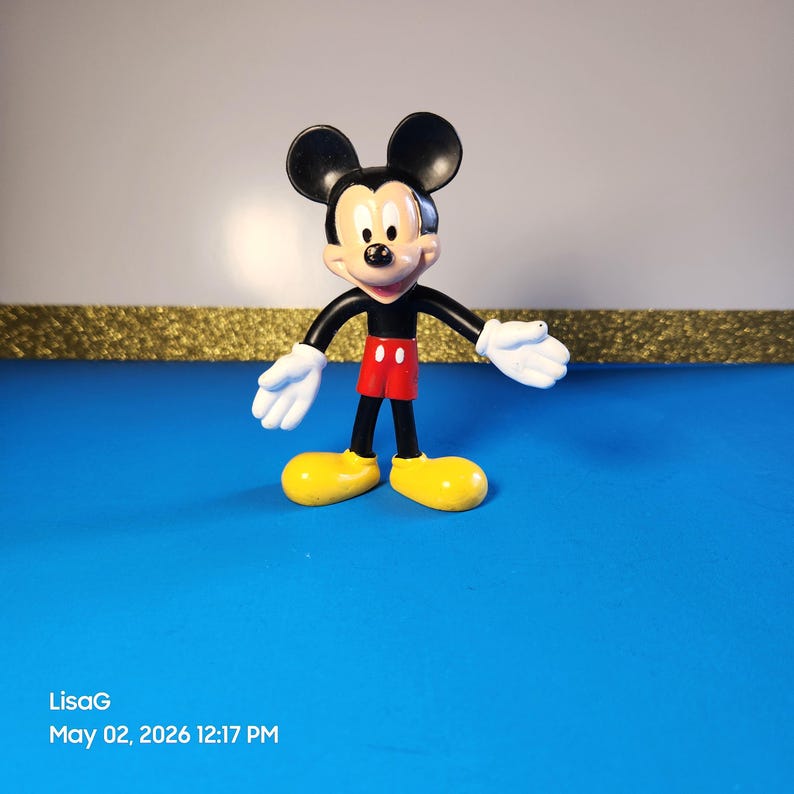 May include: A Mickey Mouse figurine with black ears, a black body, red shorts, yellow shoes, and white gloves. The figure is standing on a blue surface. The figurine is a classic Disney character.