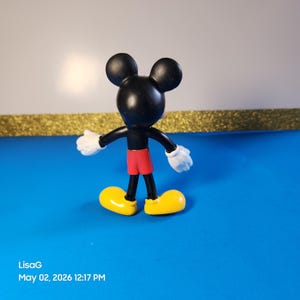 May include: A Mickey Mouse figurine, featuring a black head and body, red shorts, yellow shoes, and white gloves. The figure is posed with arms outstretched, set against a blue surface with a gold border and white background.