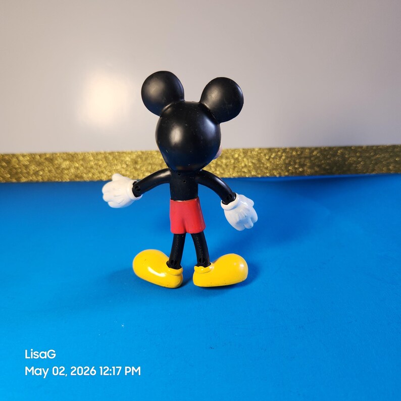 May include: A Mickey Mouse figurine, featuring a black head and body, red shorts, yellow shoes, and white gloves. The figure is posed with arms outstretched, set against a blue surface with a gold border and white background.