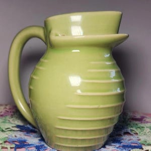 May include: A light green ceramic pitcher with a curved handle and a spout. The pitcher has horizontal ribbed detailing around the body. The pitcher is a pale green color and has a glossy finish. The pitcher is likely used for serving beverages.