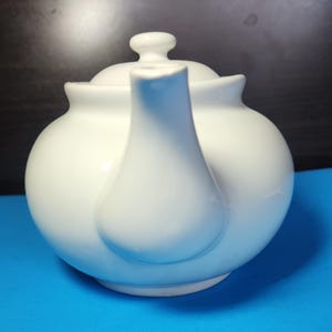 May include: A white ceramic teapot with a rounded body, a curved spout, and a small lid with a knob. The teapot is set against a blue background.