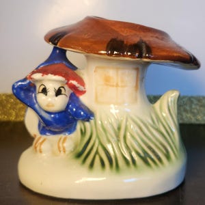 May include: A ceramic figurine featuring a small figure with a blue hat and red trim standing beside a white mushroom house. The mushroom has a brown cap and a painted window on the stem. Green grass details are painted at the base of the structure.