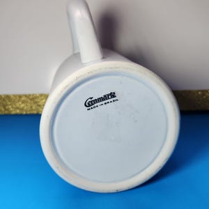 May include: A white ceramic mug with a curved handle, viewed from the bottom. The base is light blue with the text "Ceramarte MADE IN BRAZIL" in black. The mug is on a blue surface.