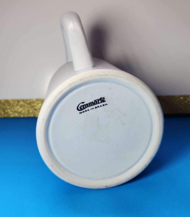May include: A white ceramic mug with a curved handle, viewed from the bottom. The base is light blue with the text "Ceramarte MADE IN BRAZIL" in black. The mug is on a blue surface.