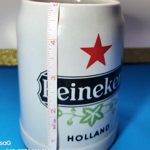 May include: White ceramic beer stein with the Heineken logo, a red star, and green accents. The stein is approximately 5 inches tall. The word "HOLLAND" is printed at the bottom.