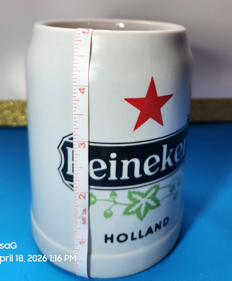 May include: White ceramic beer stein with the Heineken logo, a red star, and green accents. The stein is approximately 5 inches tall. The word "HOLLAND" is printed at the bottom.