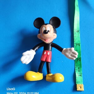 May include: A Mickey Mouse figurine with black ears, a black body, red shorts, yellow shoes, and white gloves. A green measuring tape is next to the figurine. The figurine is approximately 3 inches tall.